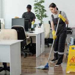 Young black man wiping computer monitors while Caucasian woman in coveralls and yellow gloves with mop cleaning floor in office